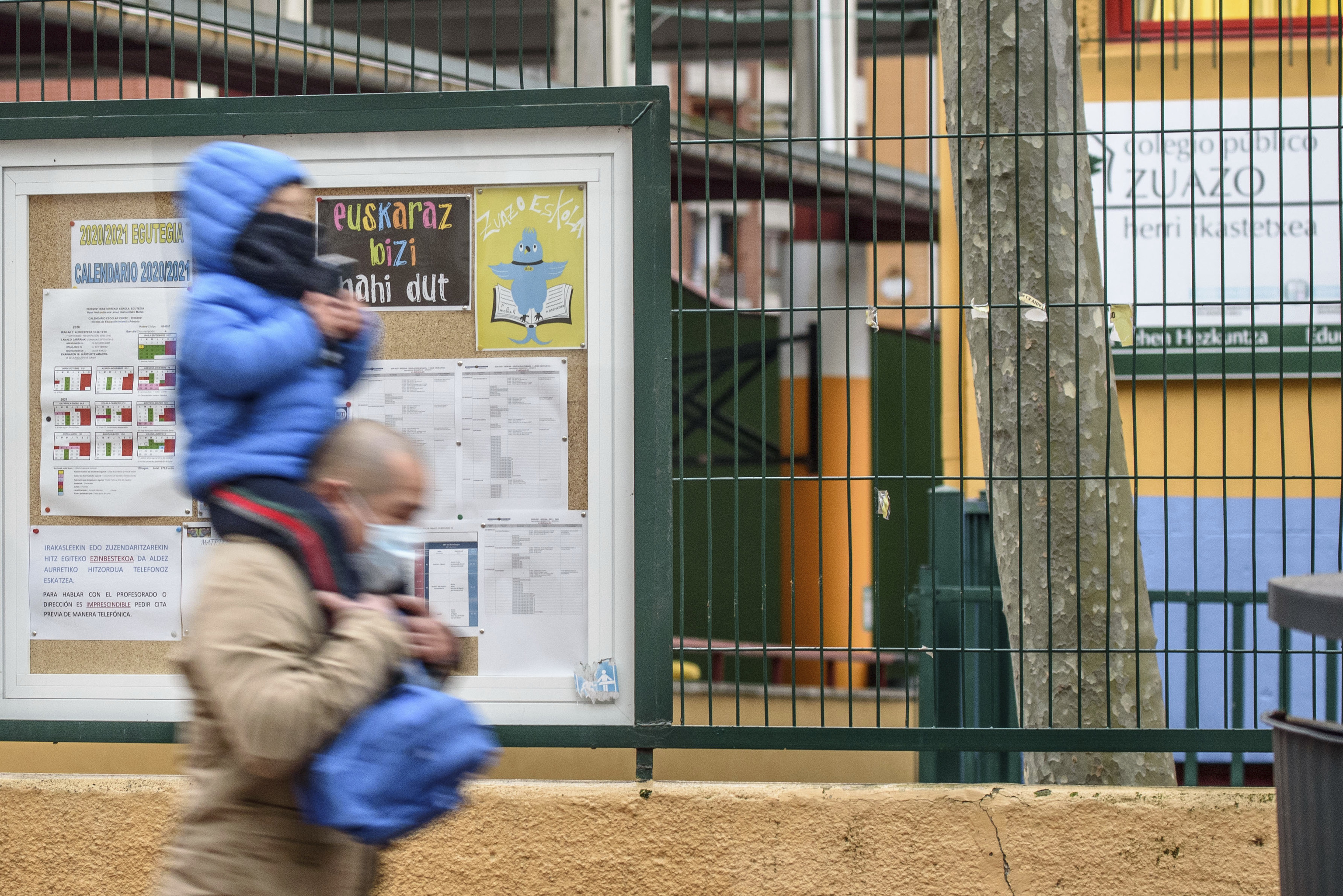 Un père porte son fils sur ses épaules devant une école avec des affiches en basque.