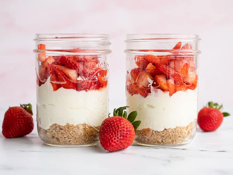 Deux pots de cheesecake aux fraises sans cuisson côte à côte avec des fraises entières sur le côté
