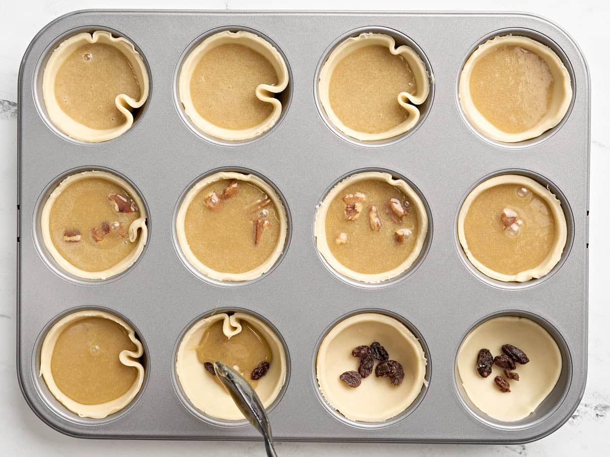 Une cuillère ajoutant la garniture pour tarte au beurre aux croûtes à tarte dans un moule à cupcake, certaines avec des pacanes et d'autres avec des raisins secs.