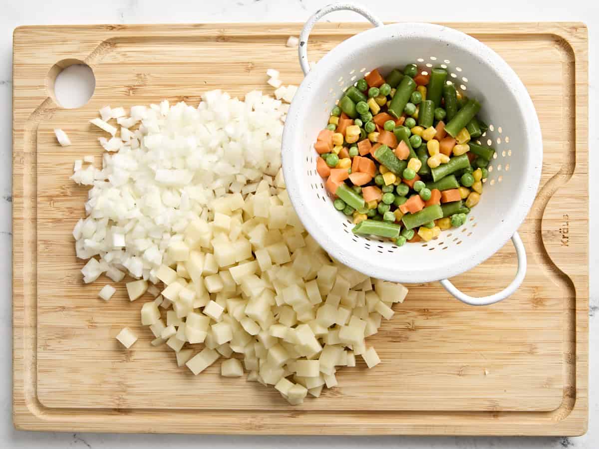 Oignon coupé en dés, pomme de terre coupée en dés et légumes surgelés sur une planche à découper en bois.