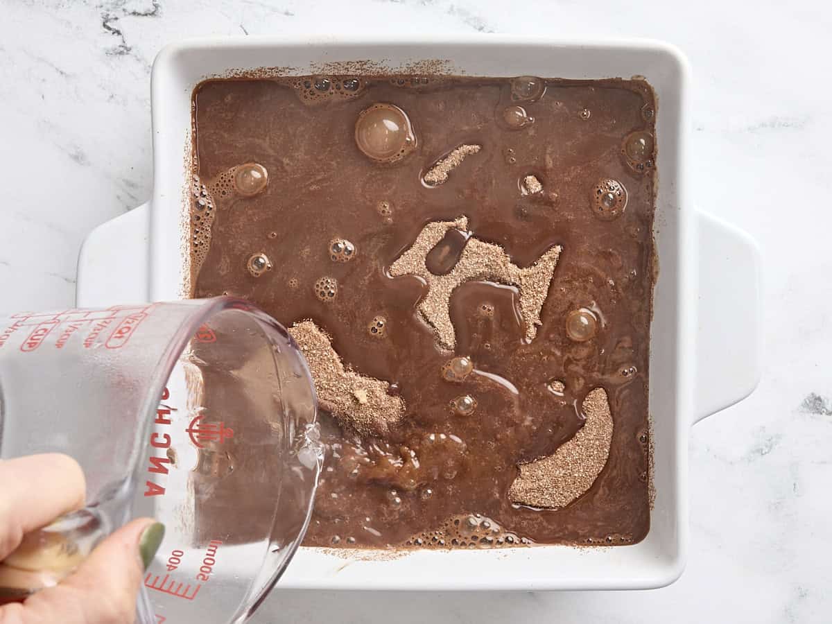 Une main versant de l'eau bouillante sur la pâte à gâteau au pouding au chocolat dans un moule à gâteau.