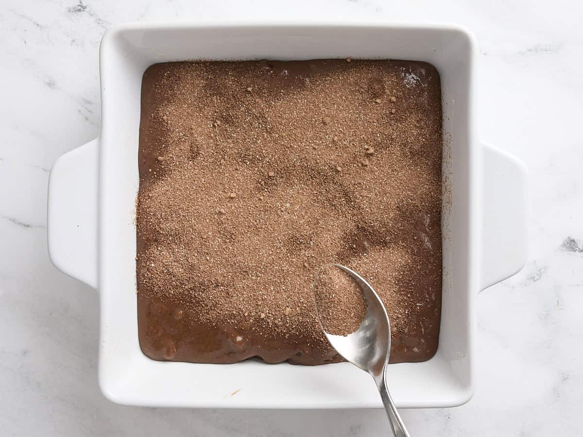 Une cuillère saupoudrant le mélange de sucre de cacao sur la pâte à gâteau au pudding au chocolat dans un moule à gâteau.