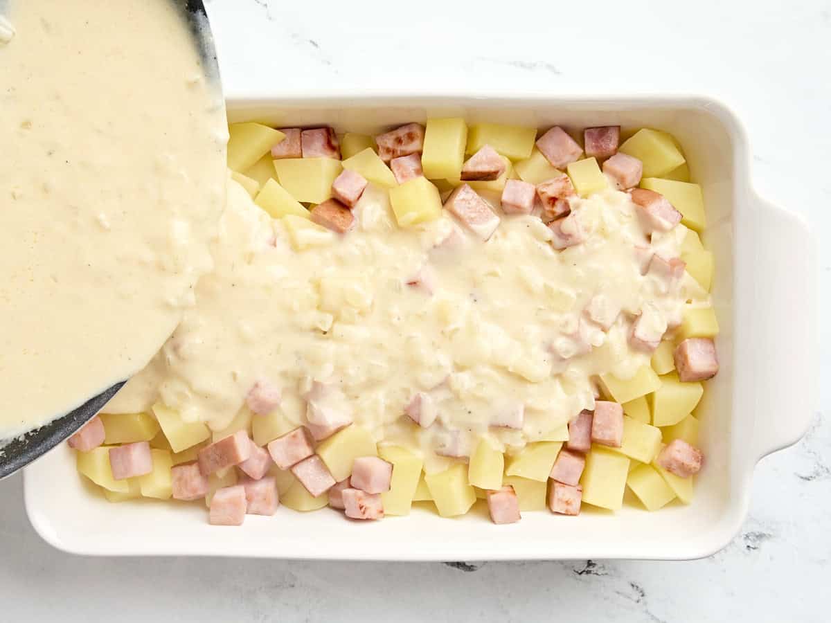 Sauce au fromage versée sur des pommes de terre en dés et un steak de jambon dans une cocotte.