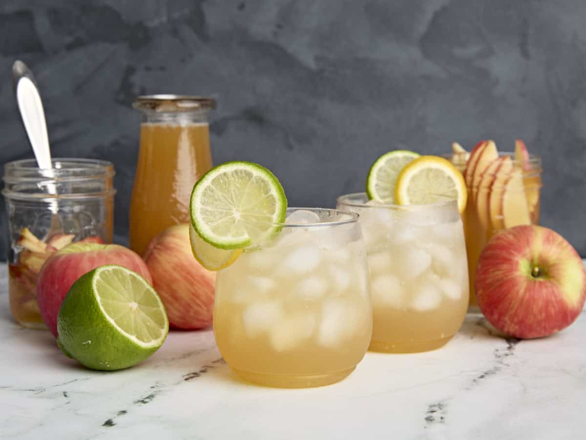 Deux cocktails sans alcool au cidre de pomme dans des verres, ganusgés de tranches de citron et de citron vert, à côté de bouteilles et de pots d'arbustes au cidre de pomme, de pommes et d'un demi citron vert.
