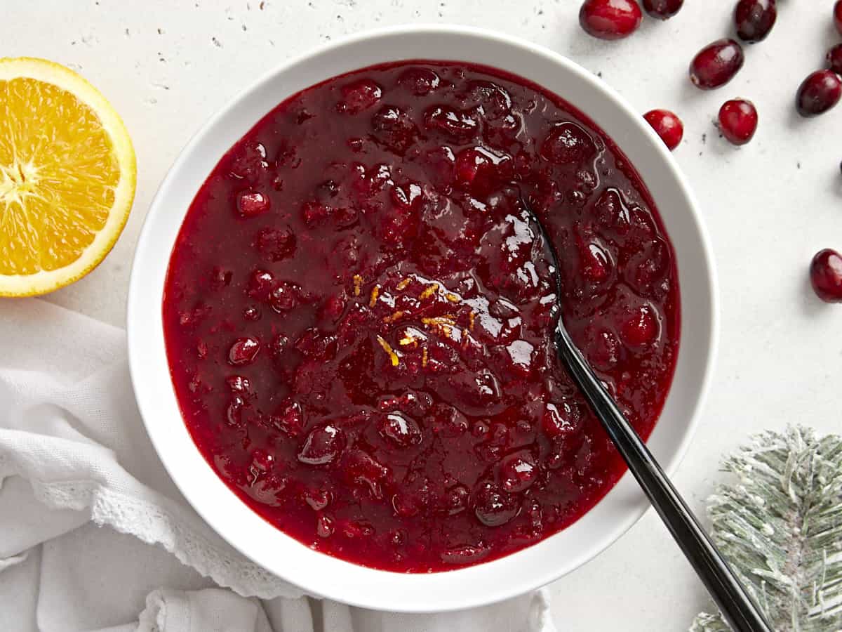 Cette simple sauce aux canneberges classique ne nécessite que trois ingrédients mais a suffisamment de saveur pour ajouter du piquant à votre repas des Fêtes. Budgetbytes.com Vue aérienne de la sauce aux canneberges dans un bol avec du zeste d'orange et une cuillère.