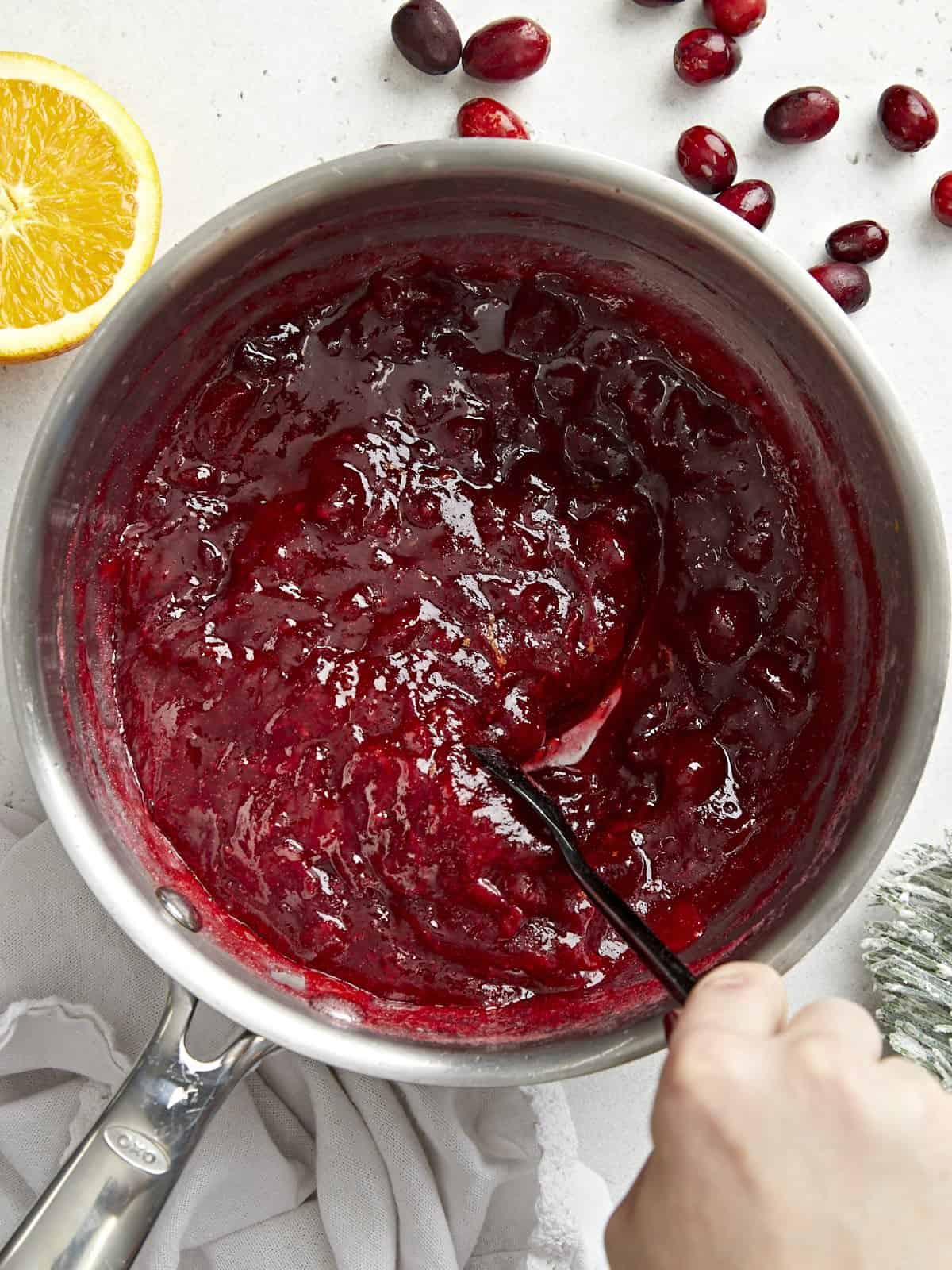 Cette simple sauce aux canneberges classique ne nécessite que trois ingrédients mais a suffisamment de saveur pour ajouter du piquant à votre repas des Fêtes. Budgetbytes.com Vue aérienne d'un pot rempli de sauce aux canneberges maison en cours d'agitation.