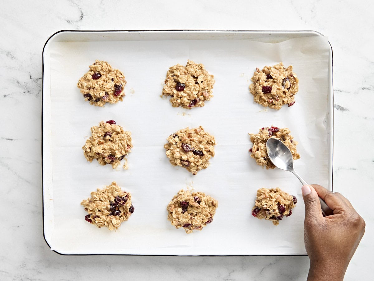 Portions de pâte à biscuits pour le petit-déjeuner sur une plaque à pâtisserie tapissée de papier sulfurisé, avec une cuillère en appuyant sur les portions pour les aplatir.