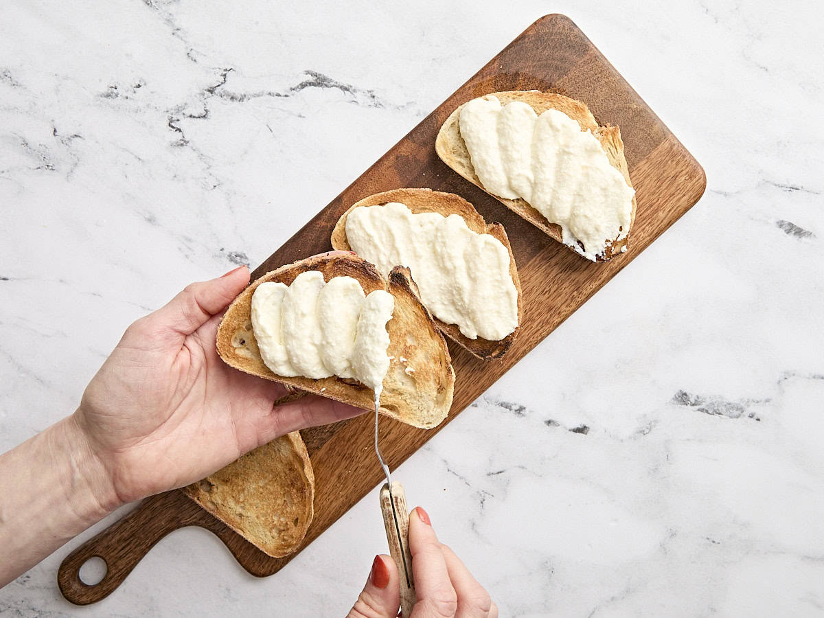 Une main étalant de la ricotta fouettée sur des tranches de pain grillé au levain.