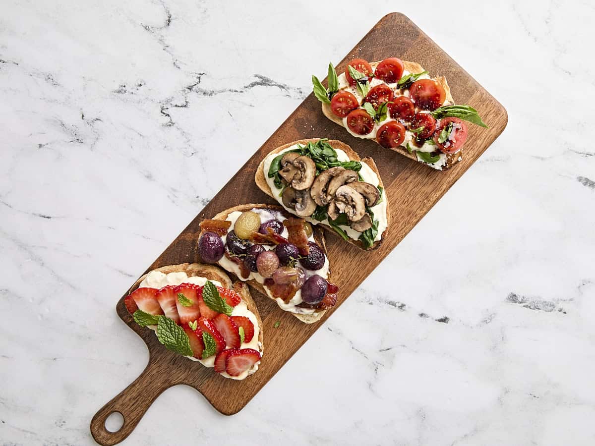Toast à la ricotta de 4 façons sur un plateau de service en bois, l'une garnie de fraises et de menthe, la suivante de raisins rôtis et de bacon, une autre de tomates grillées et de basilic, et la dernière de champignons et d'épinards fanés.