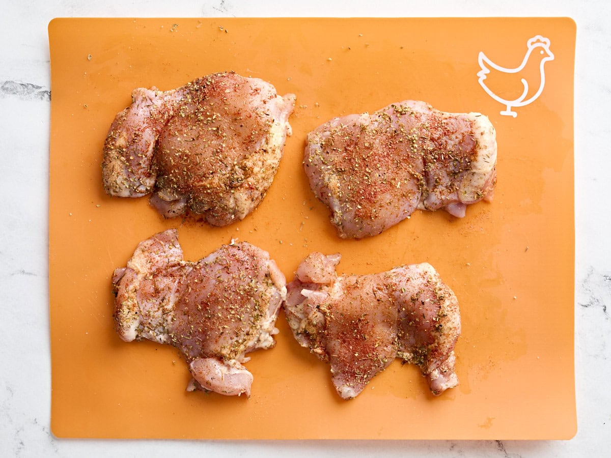 Cuisses de poulet assaisonnées d'un mélange d'assaisonnement grec sur un tapis de préparation de viande.