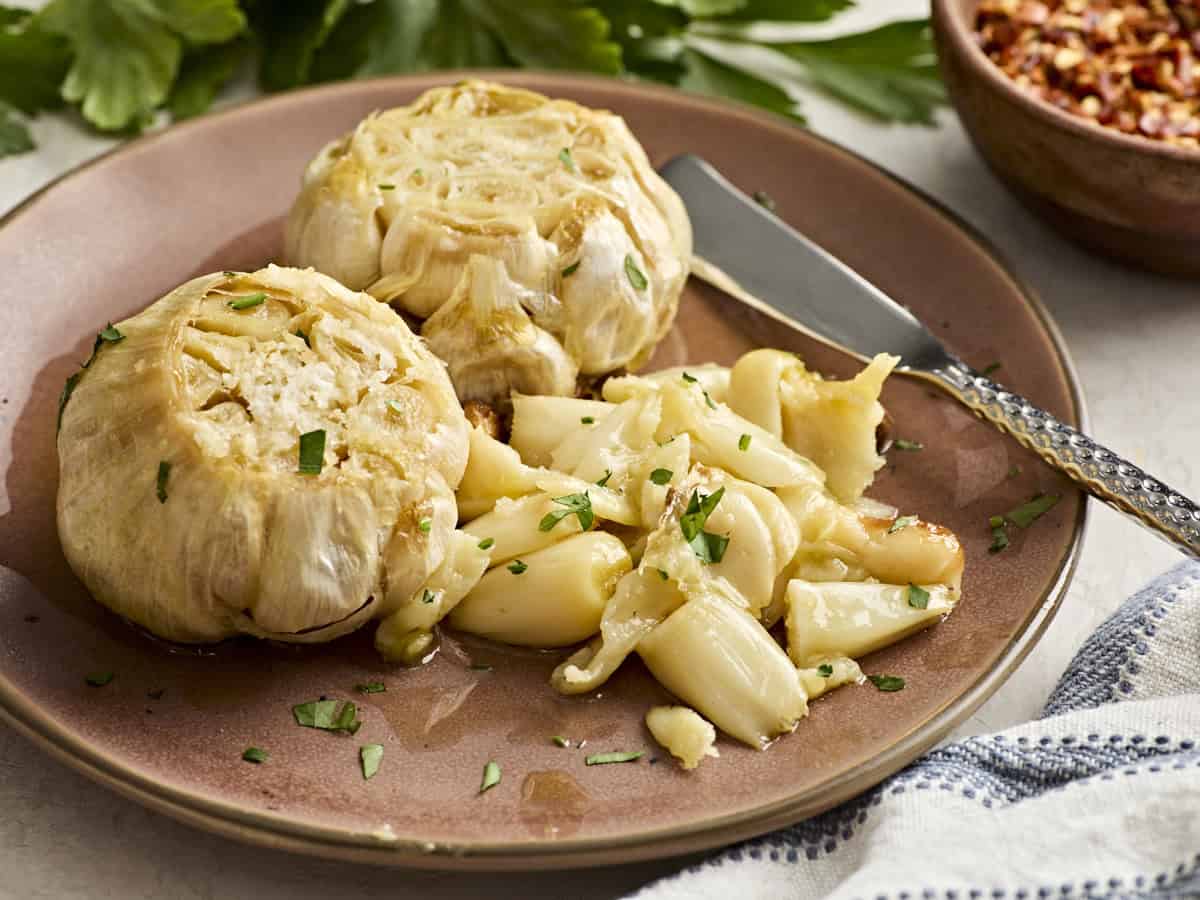 Vue latérale de deux têtes d'ail rôties sur une assiette avec un couteau et des gousses d'ail.