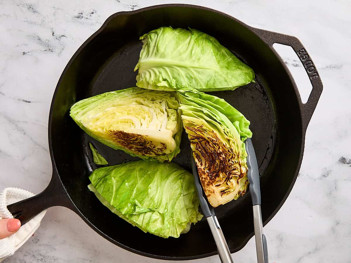 Des pinces de cuisine retournant sur les steaks de chou dans une poêle en fonte.