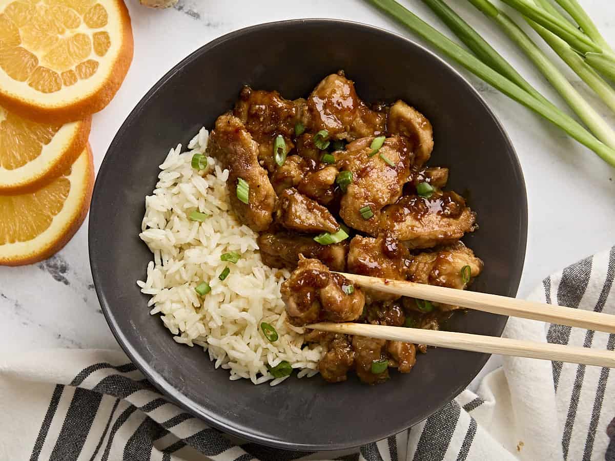 Ce poulet orange facile cuit rapidement dans une poêle (pas de friture!) Et utilise uniquement des ingrédients réels et frais. C'est le faux faux-net parfait. Budgetbytes.com Vue aérienne du poulet orange dans un bol avec du riz, avec des baguettes prenant un morceau de poulet.