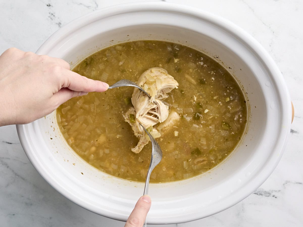 Mains utilisant deux fourches pour déchiqueter une poitrine de poulet dans une mijoteuse.