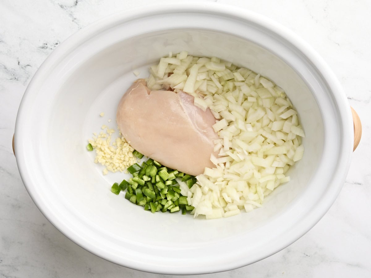 Une poitrine de poulet, de l'ail haché, de l'oignon en dés et du jalapeno dans une mijoteuse.