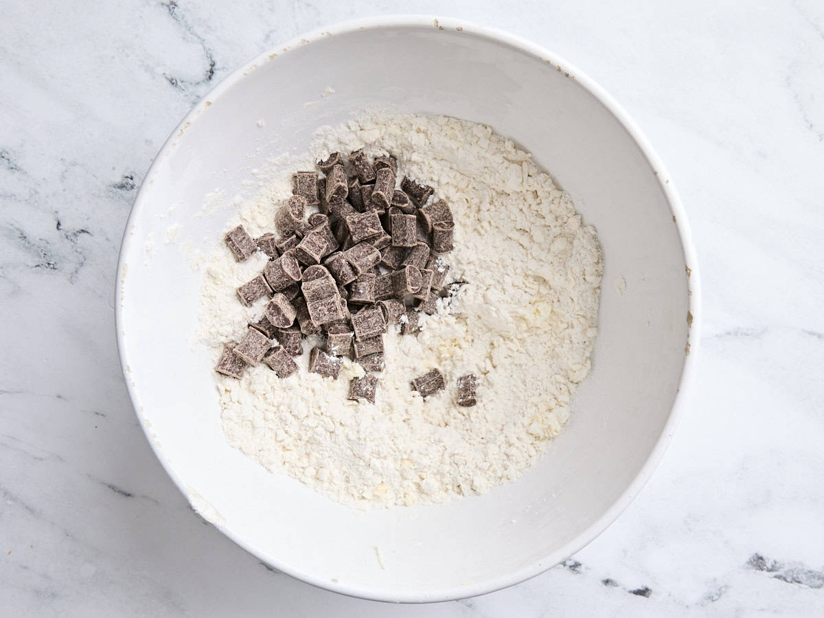 Des morceaux de chocolat ajoutés à un mélange à quatre et au beurre dans un bol à mélanger blanc.