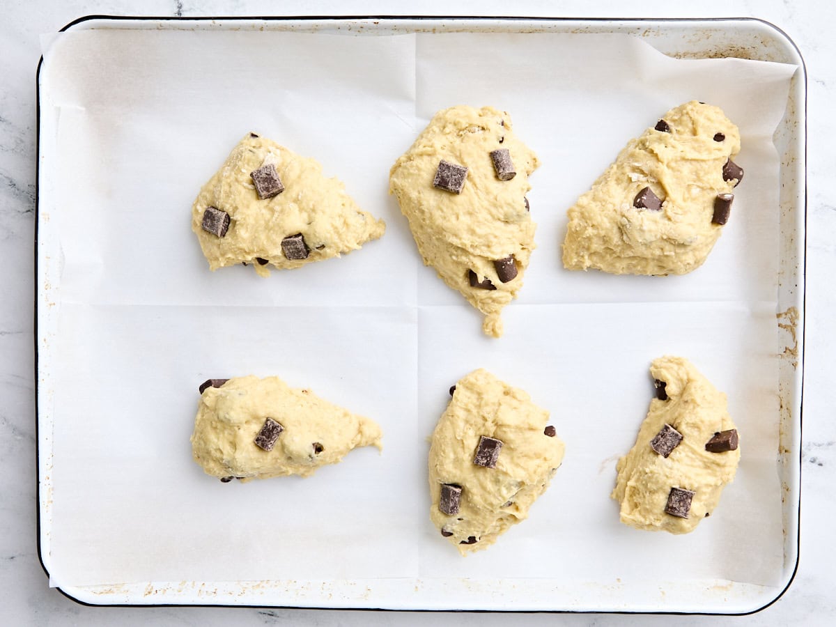 Des scones aux pépites de chocolat à six formes sur une plaque à pâtisserie doublée de parchemin prête à être cuite.