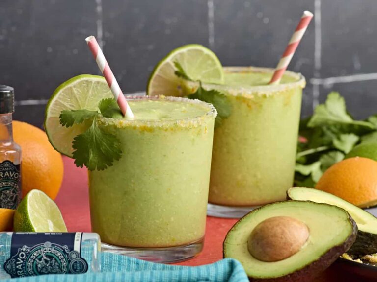 Side view of two glasses of avocado margarita, garnished with lime slices and fresh cilantro.