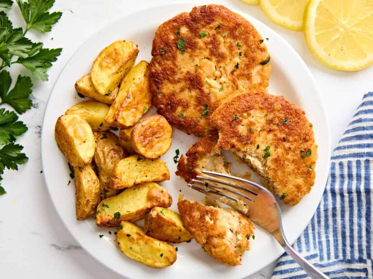 Two salmon patties on a plate, with one sliced in half by a fork, with a side of air fried potatoes.
