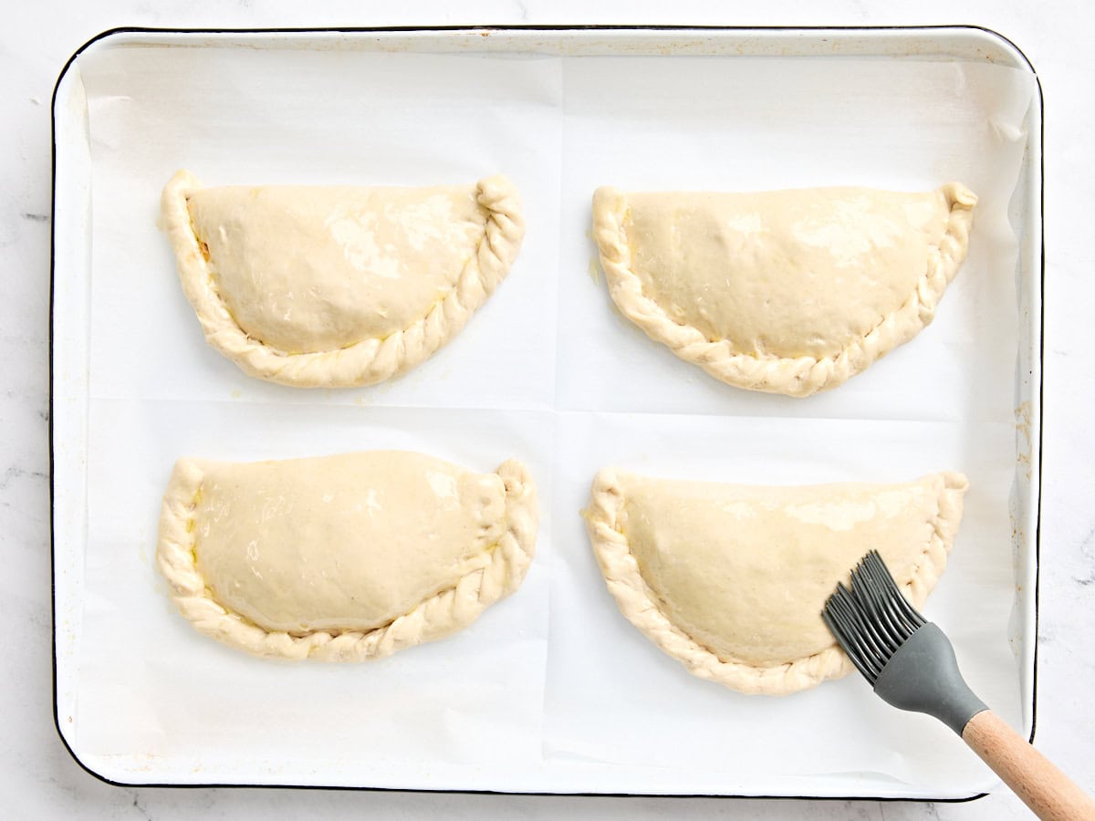 Quatre calzones remplies sur une plaque à pâtisserie doublée de parchemin sont brossées d'huile d'olive avant la cuisson.