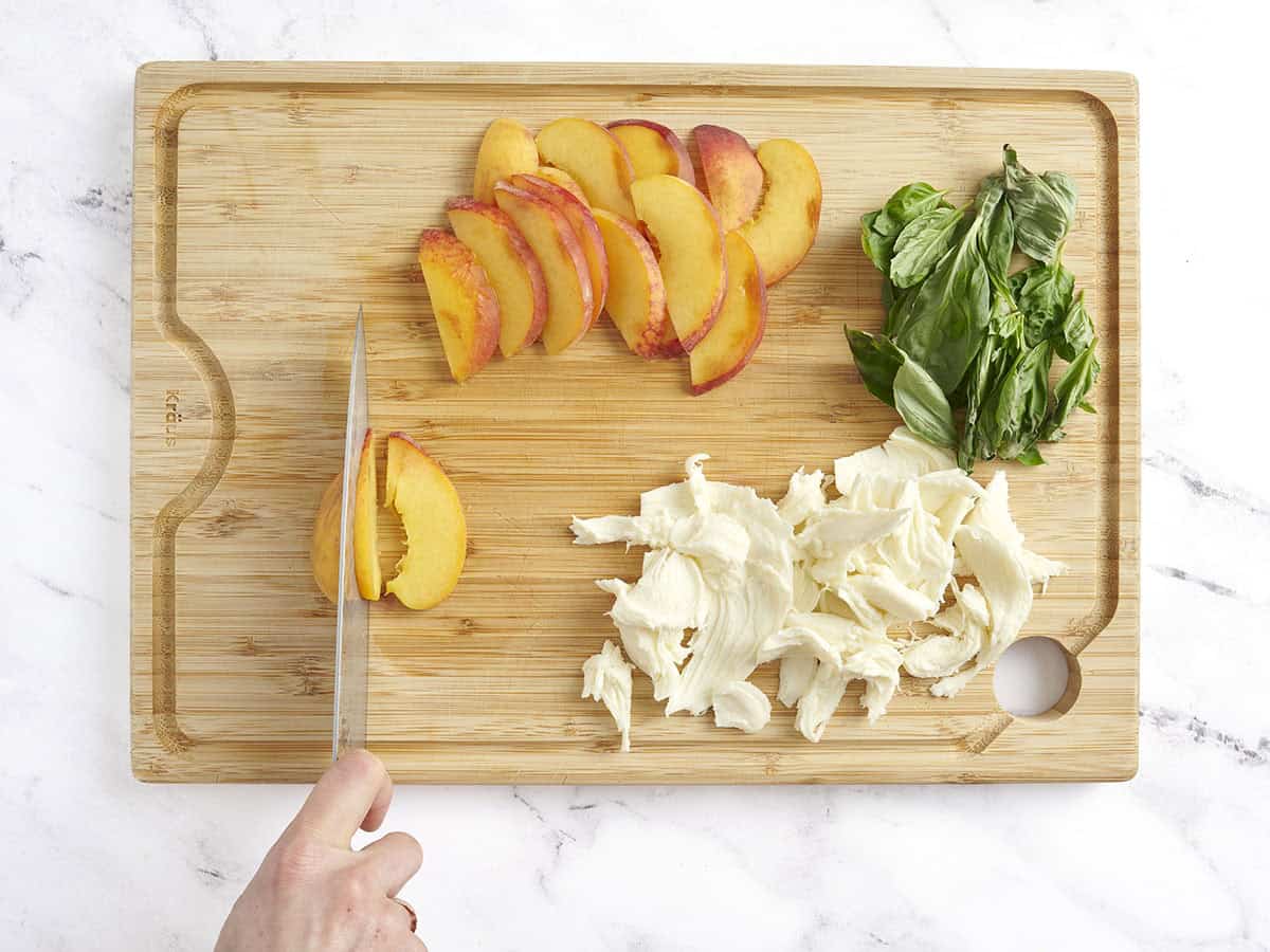 Un couteau tranchant des pêches sur une planche à découper en bois à côté de la mozzarella fraîche et des feuilles de basilic,