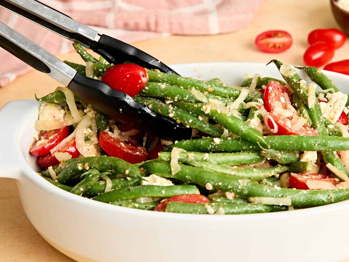 Vue latérale des pinces de cuisine prenant une salade de haricots verts dans un plat.