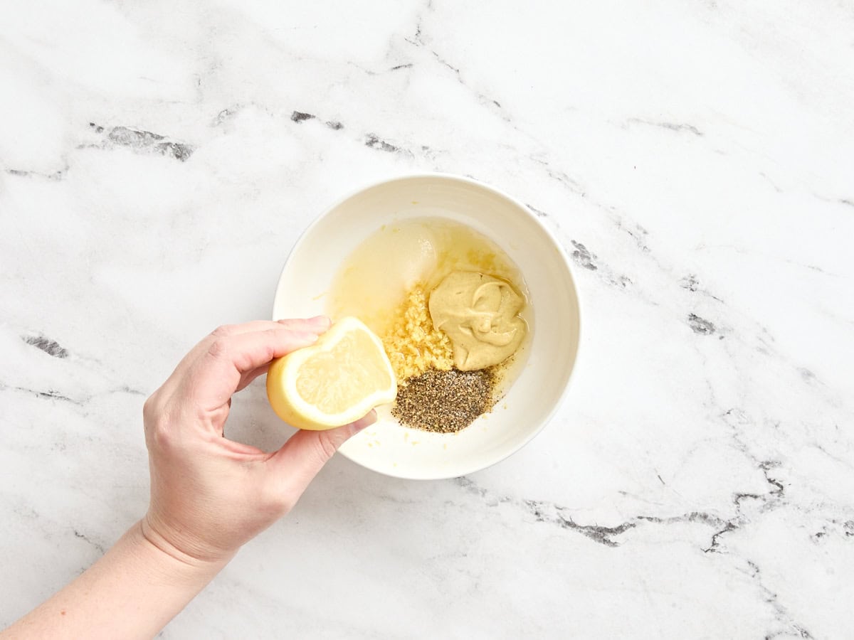 Une main pressant un demi-citron dans un bol de vinaigre de cidre de pomme, de moutarde, de sucre et d'assaisonnements.