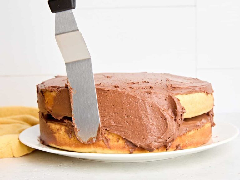 Side view of a vanilla cake being spread with chocolate buttercream frosting.