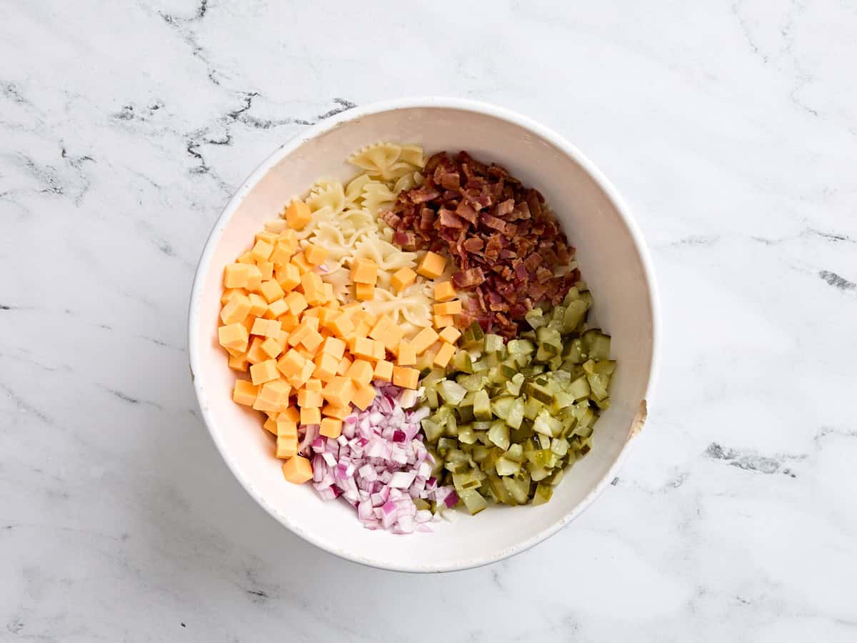 Pâtes en noeud papillon cuites, cheddar en cubes, bacon cuit et émietté, cornichons d'aneth en dés. et l'oignon rouge en dés dans un bol.