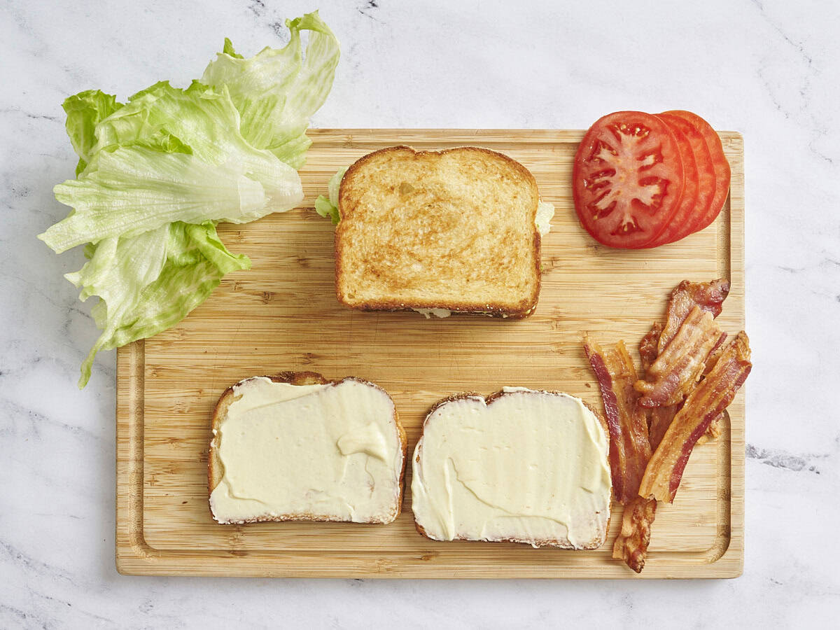 Vue aérienne de deux tranches de pain grillées garnies de mayo, de bacon cuit, de laitue, de tomate tranchée et d'un sandwich BLT terminé