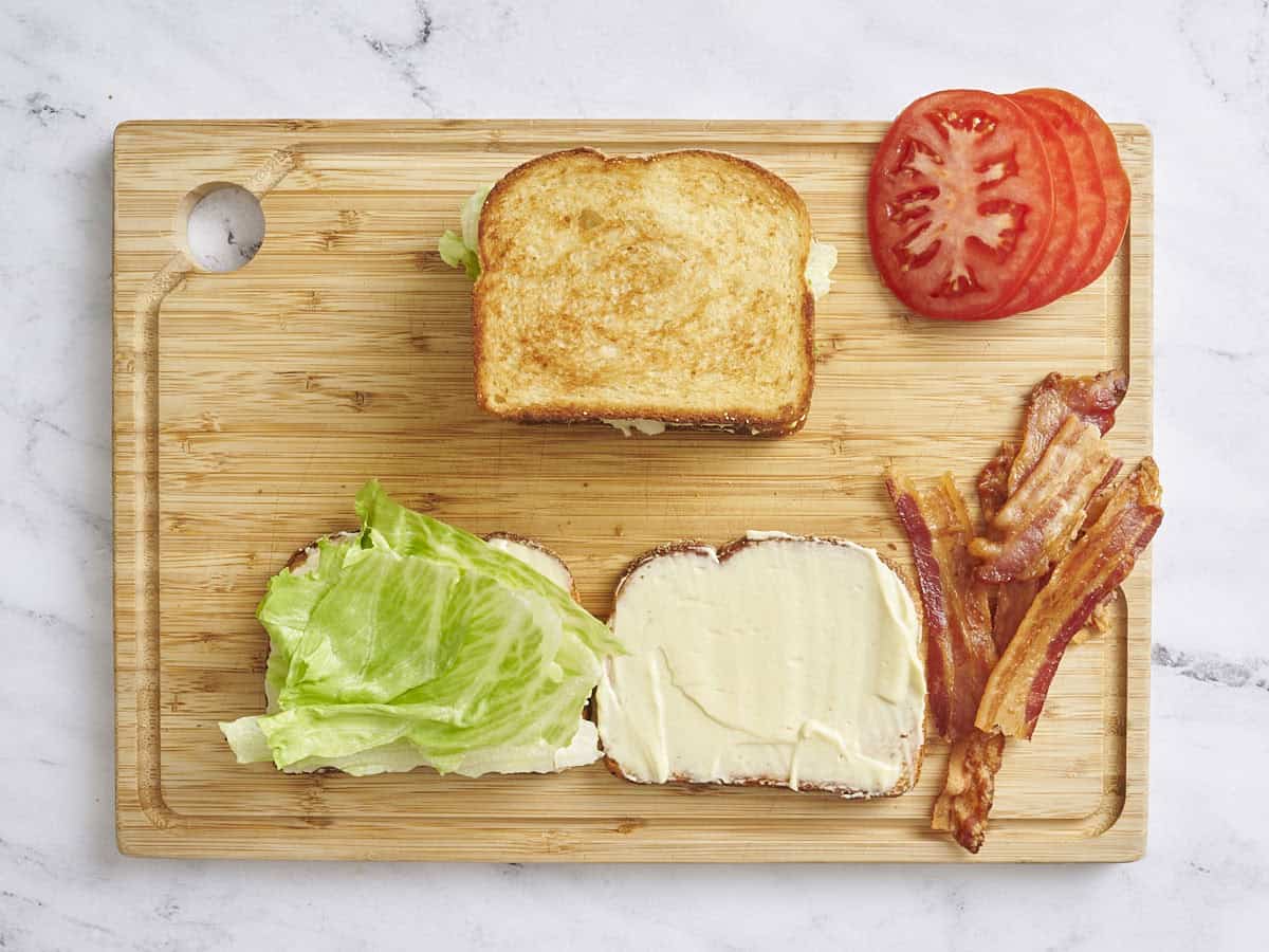 Vue aérienne d'un sandwich BLT assemblé sur une planche à découper en bois, avec des tomates tranchées, du bacon cuit et deux tranches de pain ouvertes à côté avec une tranche garnie de laitue.