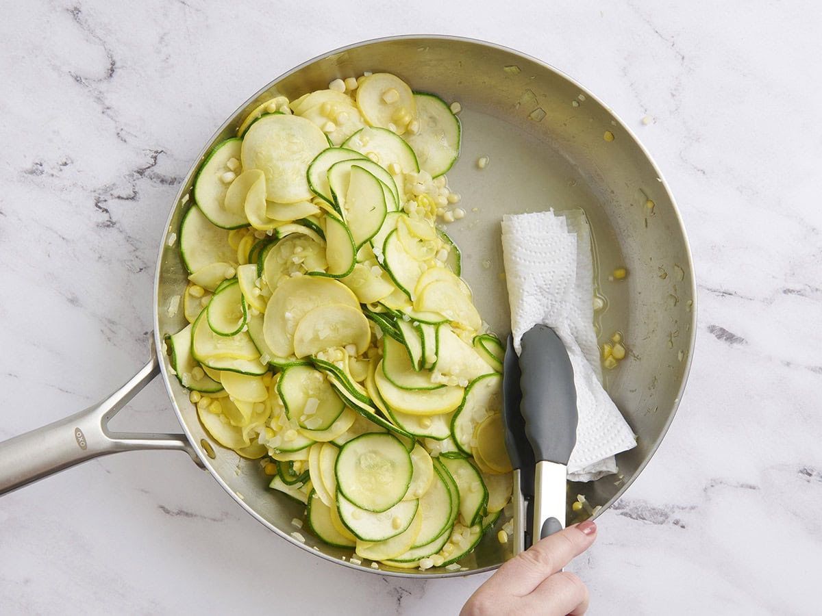 Des pinces de cuisine absorbant le liquide des courgettes sautées et de la courge jaune dans une poêle.
