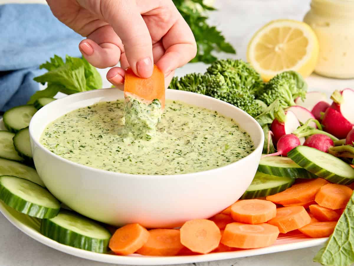 Side view of a bowl of green goddess salad dressing on a platter of fresh vegetables with a hand dipping a carrot in.