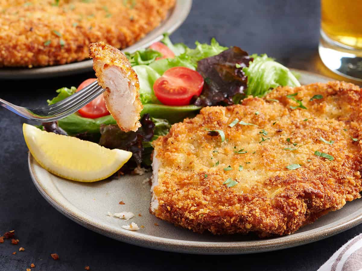 Vue latérale d'un schnitzel de poulet dans une assiette avec une fourchette prenant une tranche.