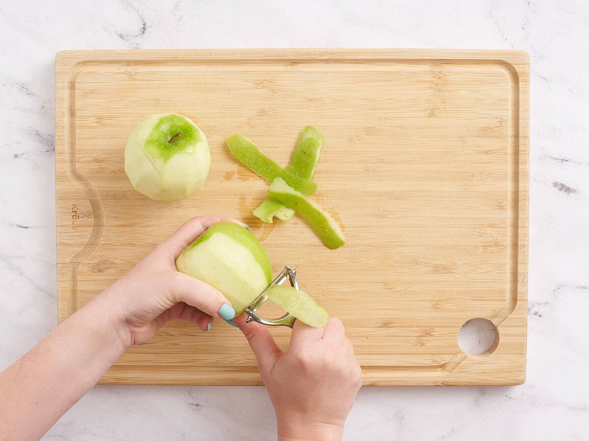 Une main utilisant un éplucheur de légumes pour peler une pomme verte.