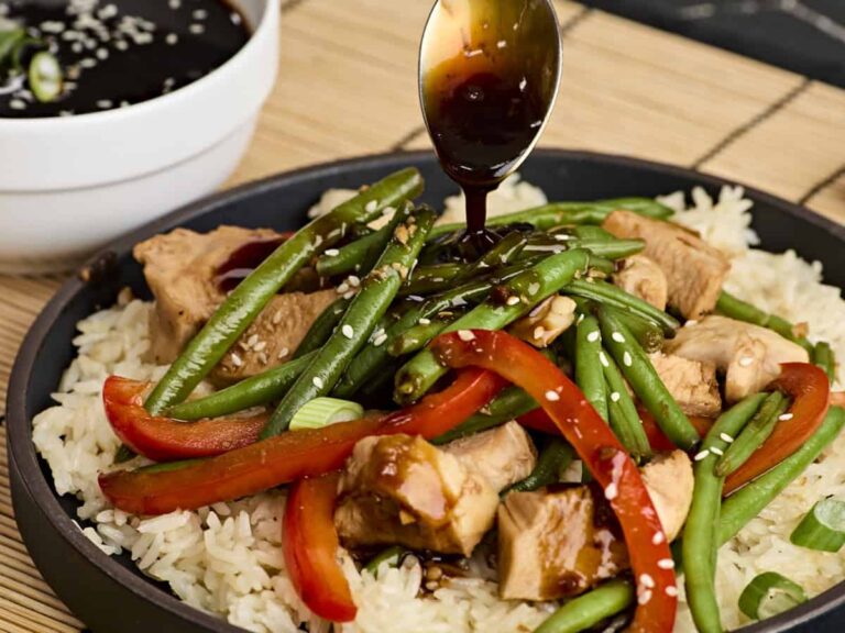 A spoon drizzling teriyaki sauce over a plate of rice, chicken, green beans, and red bell peppers.