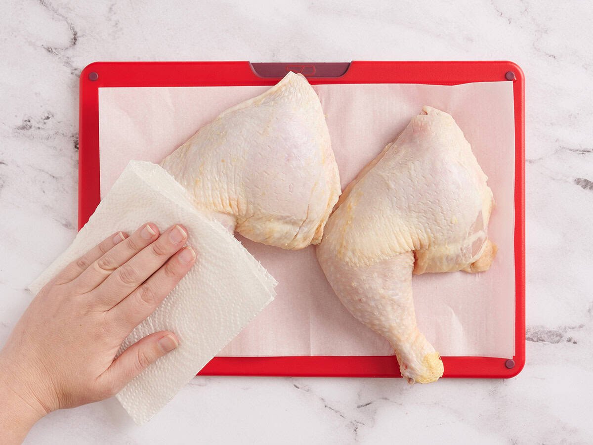 Une main tapotant une cuisse de poulet pour la sécher avec une serviette en papier.