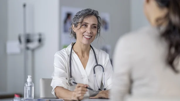 Une femme médecin mature montre l'écran de sa tablette posée sur la table devant elle alors qu'elle partage avec elle les résultats des tests de ses patients. Elle porte une blouse blanche et un stéthoscope autour du cou alors qu'elle parle avec la patiente d'origine asiatique.