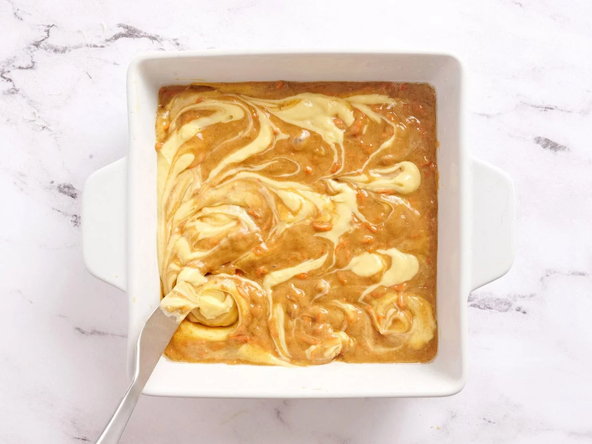 Le cheesecake tourbillonne sur des barres de gâteau aux carottes dans un plat allant au four.