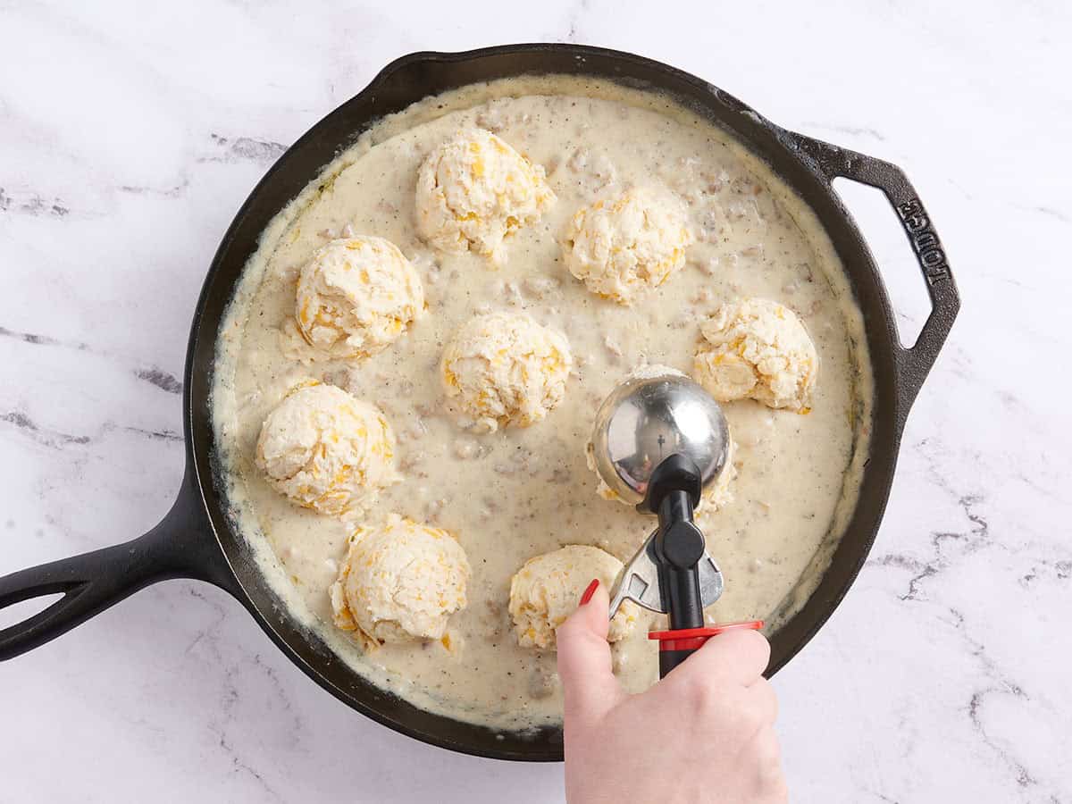 Une main utilisant une cuillère pour répartir la pâte à biscuit sur la sauce à saucisse.