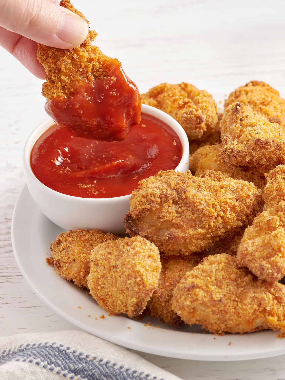 Vue latérale d'une assiette de nuggets de poulet faits maison avec une main trempant une nugget dans un pot de ketchup.