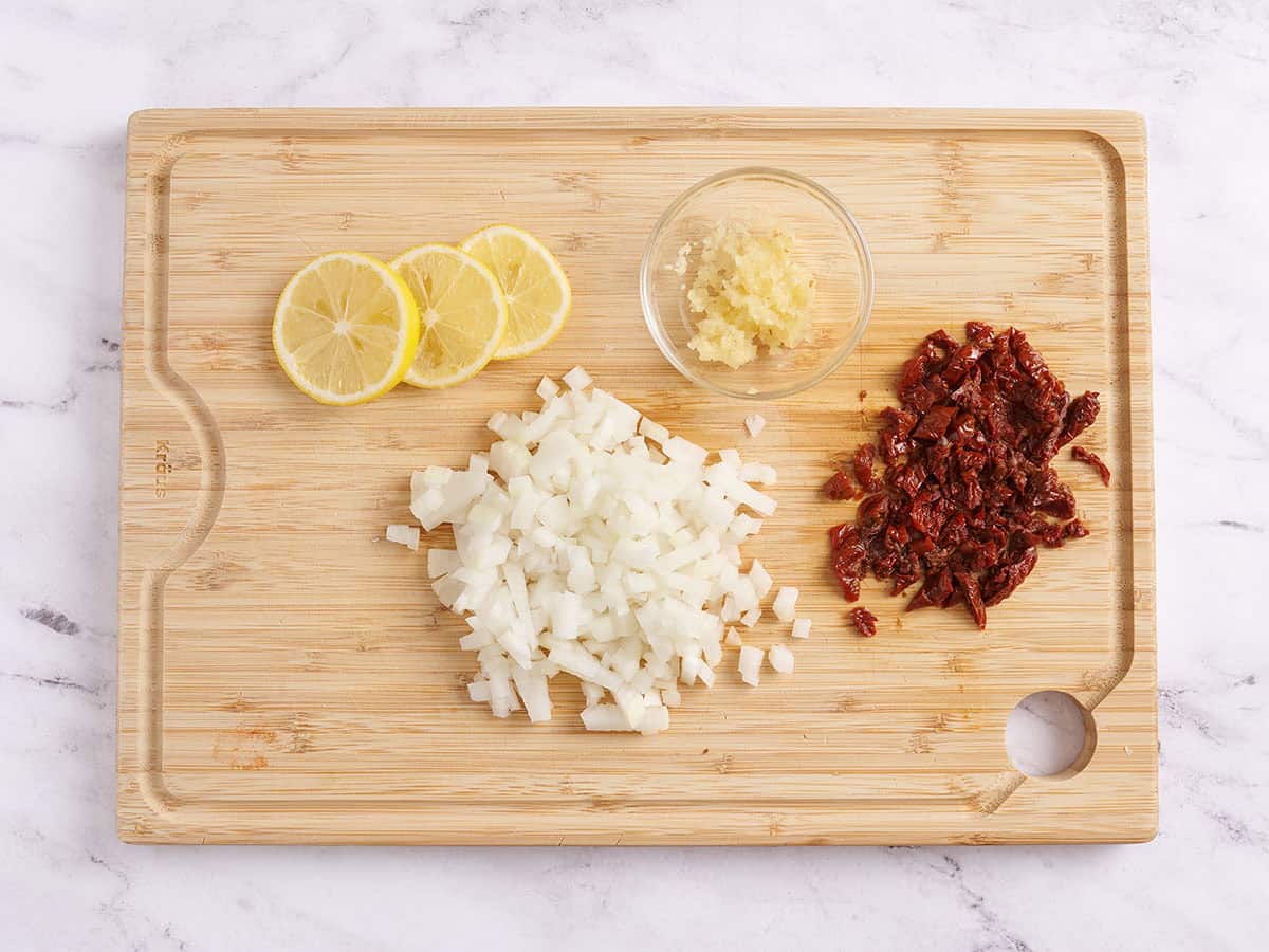 Citrons tranchés, oignons coupés en dés, tomates séchées coupées en dés et ail émincé sur une planche à découper en bois.