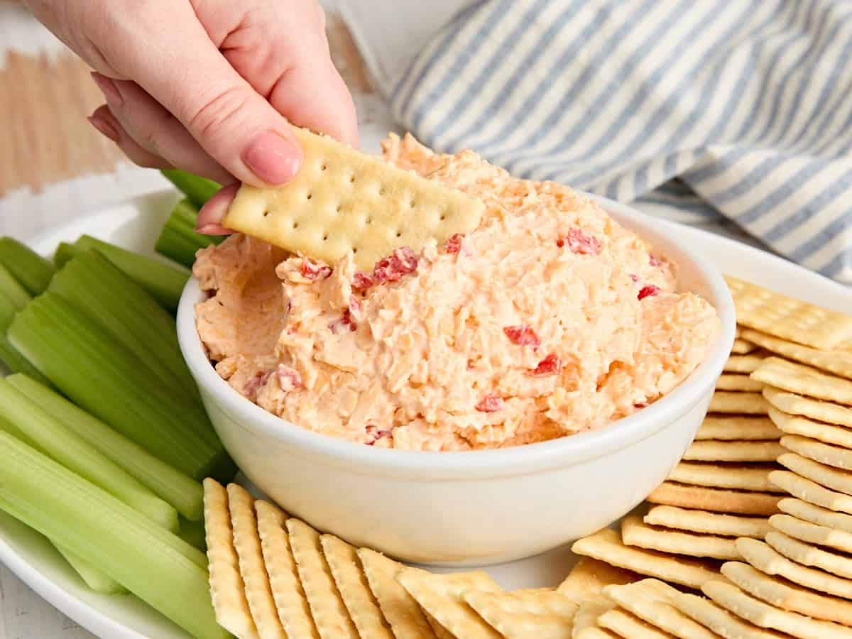 Vue latérale d'un bol de trempette au fromage au piment du sud avec une main trempant un cracker dedans.