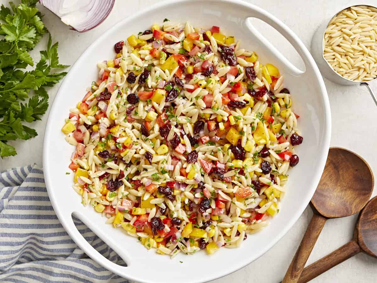 Vue aérienne de la salade de pâtes Orzo dans un bol de service blanc avec des cuillères de service en bois sur le côté.