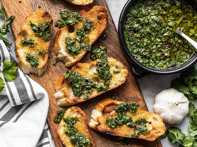 Pain français grillé en tranches sur une planche à découper arrosé de chimichurri avec un bol de sauce chimichurri sur le côté.