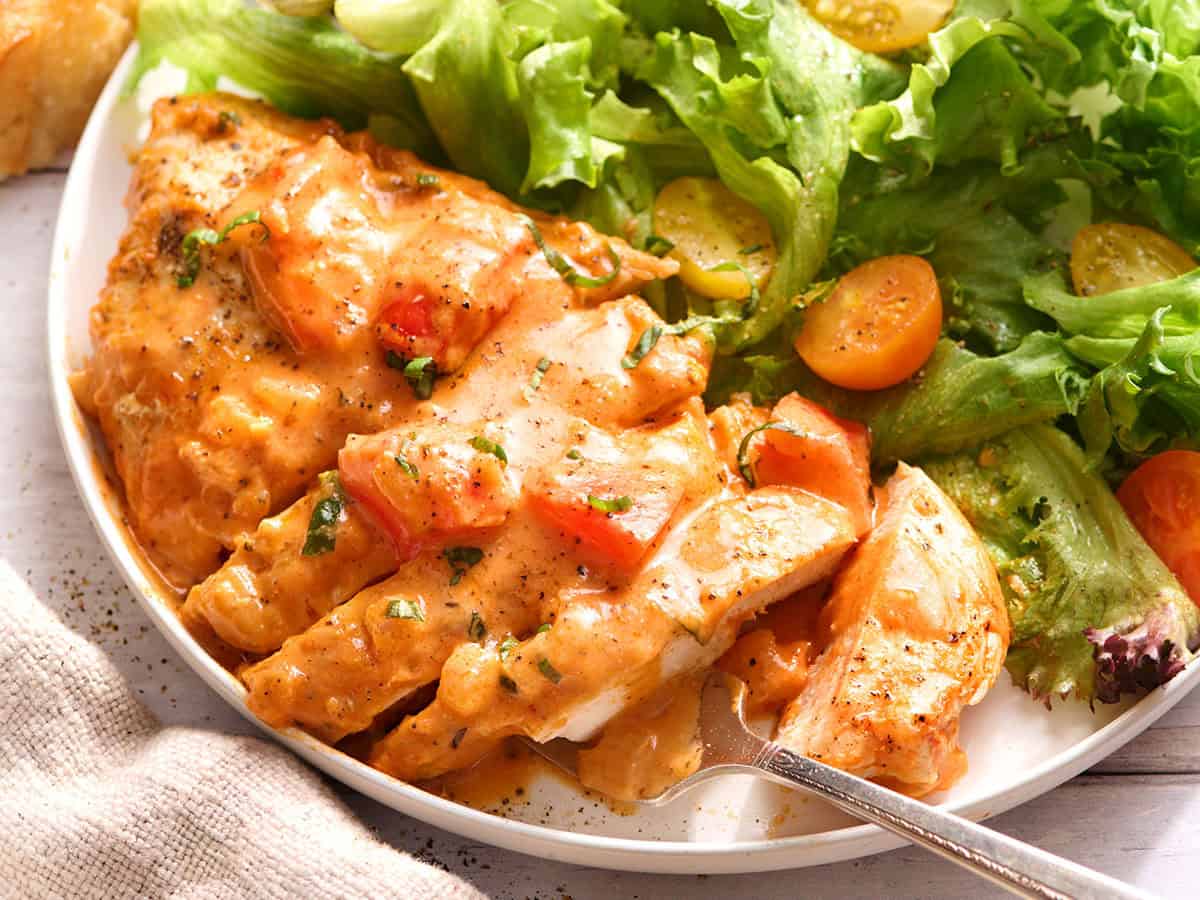 Side view of a creamy tomato chicken breast cut into slices on a plate with a green salad.