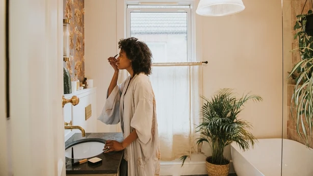 Une belle femme noire se tient dans une salle de bains élégante, vêtue d’une longue robe soyeuse et se maquille soigneusement.