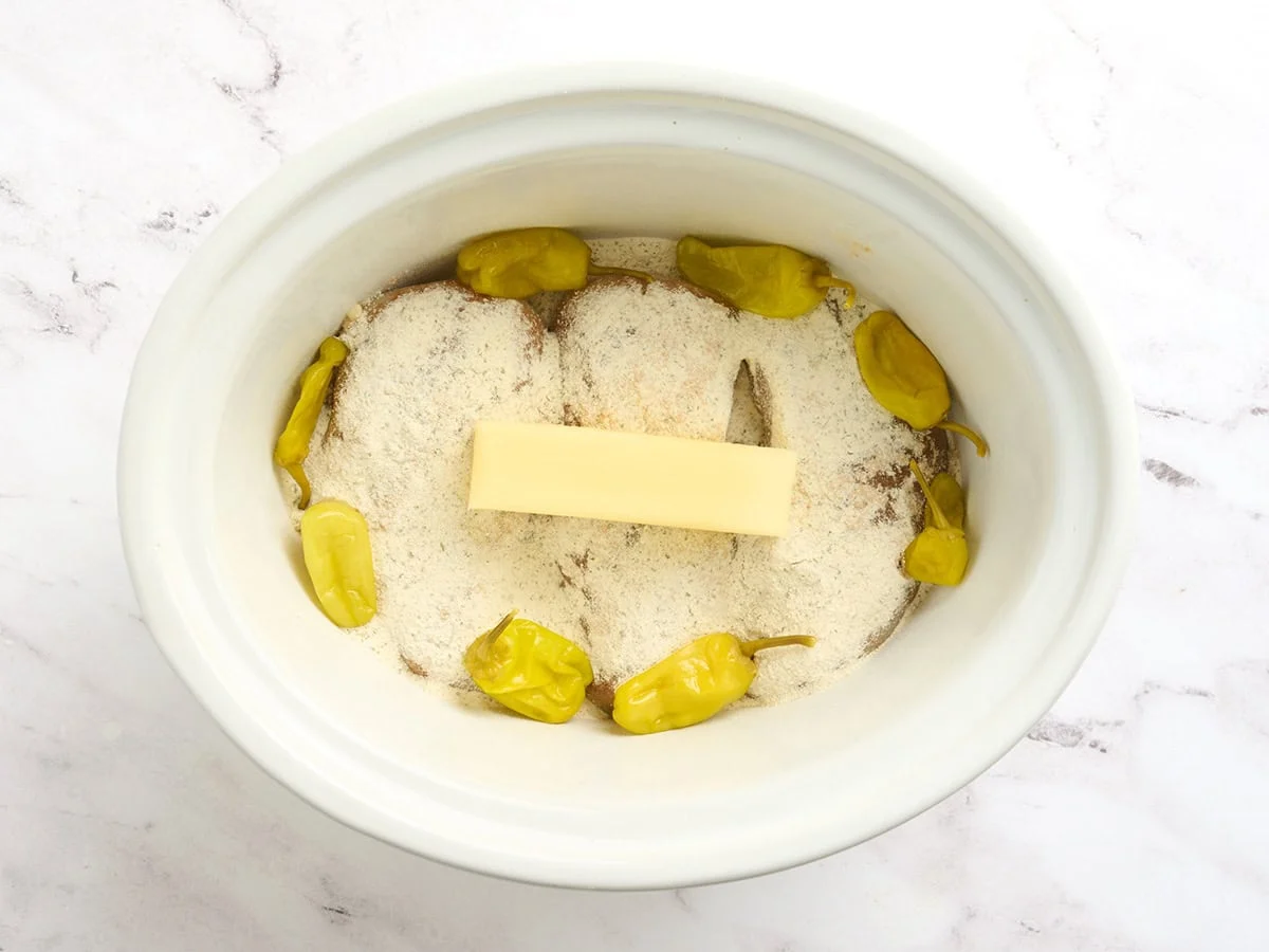 Poitrines de poulet dans une mijoteuse avec des assaisonnements, un bâton de beurre et des poivrons pepperoncini pour faire du poulet du Mississippi.