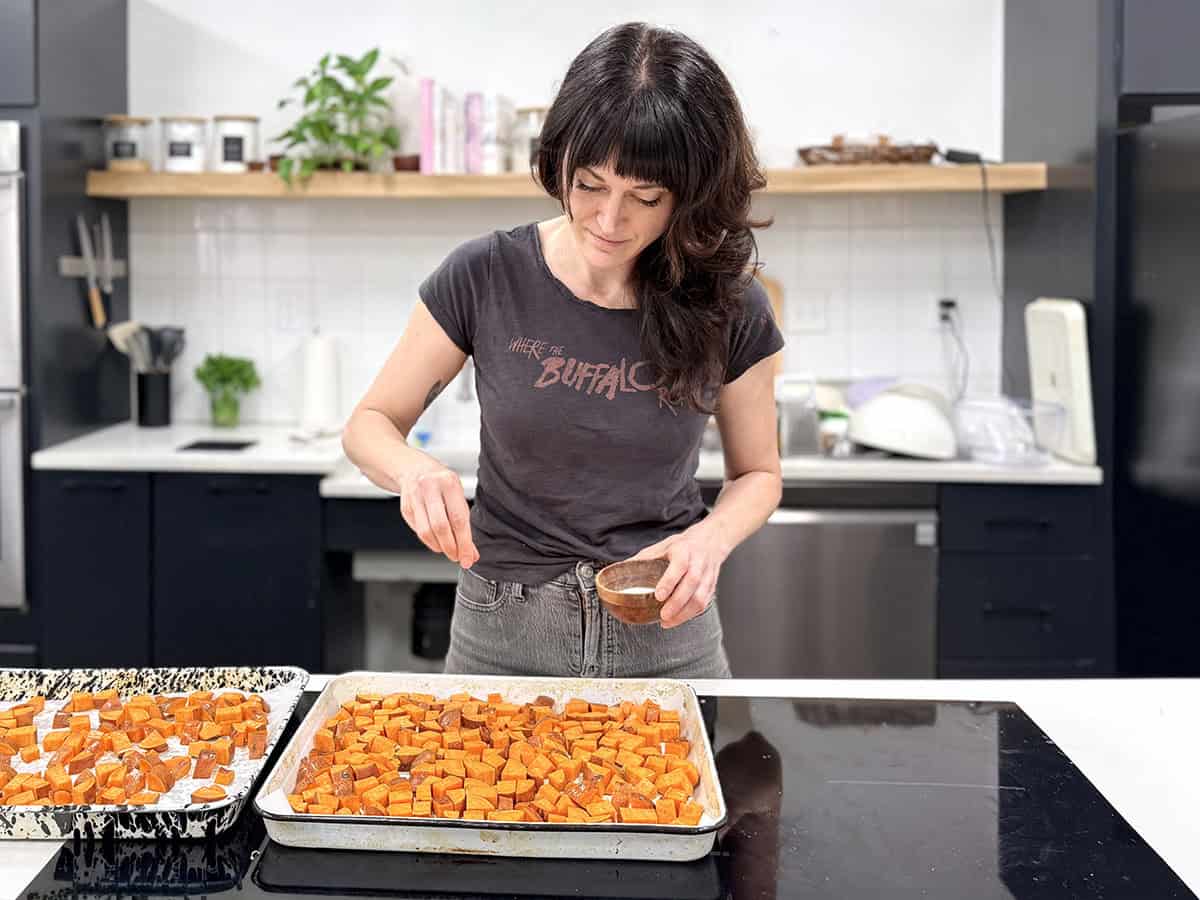 Une femme saupoudre du sel sur une plaque de patates douces coupées en dés dans une cuisine.